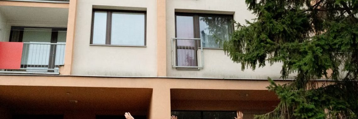 Three women waving to an elderly woman on an apartment balcony in Prague.